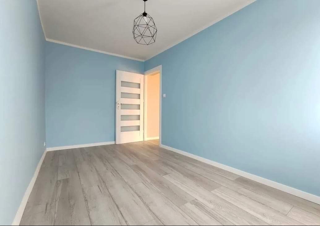 Jasne, błękitne ściany w pokoju z szarymi panelami podłogowymi i nowoczesną lampą wiszącą, białe drzwi z przeszkleniami po lewej stronie, widok na kolejne pomieszczenie.