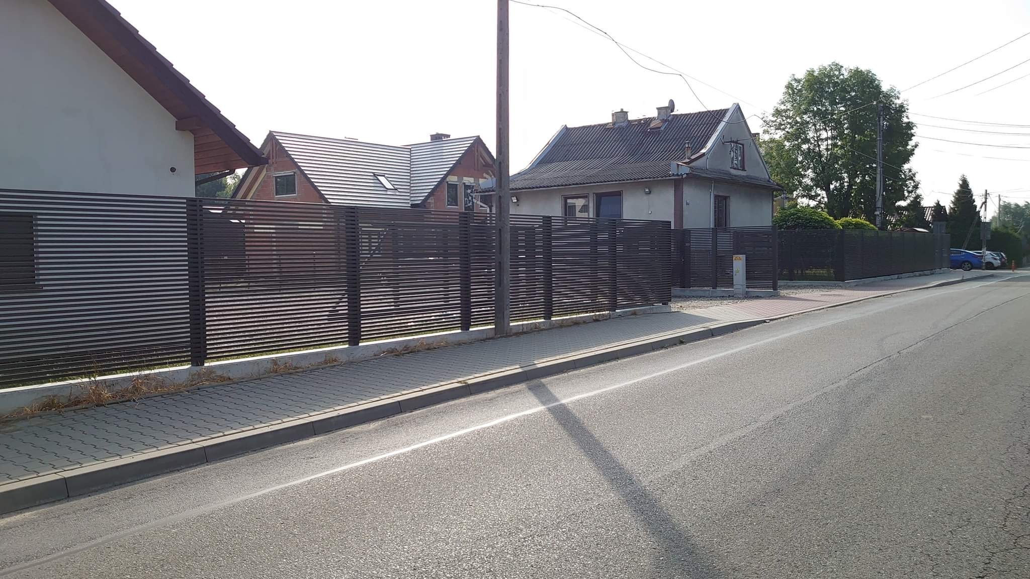 Nowoczesne ogrodzenie panelowe z poziomych listew w kolorze antracytowym, oddzielające posesje od ulicy z chodnikiem wyłożonym kostką brukową, widok z poziomu jezdni.