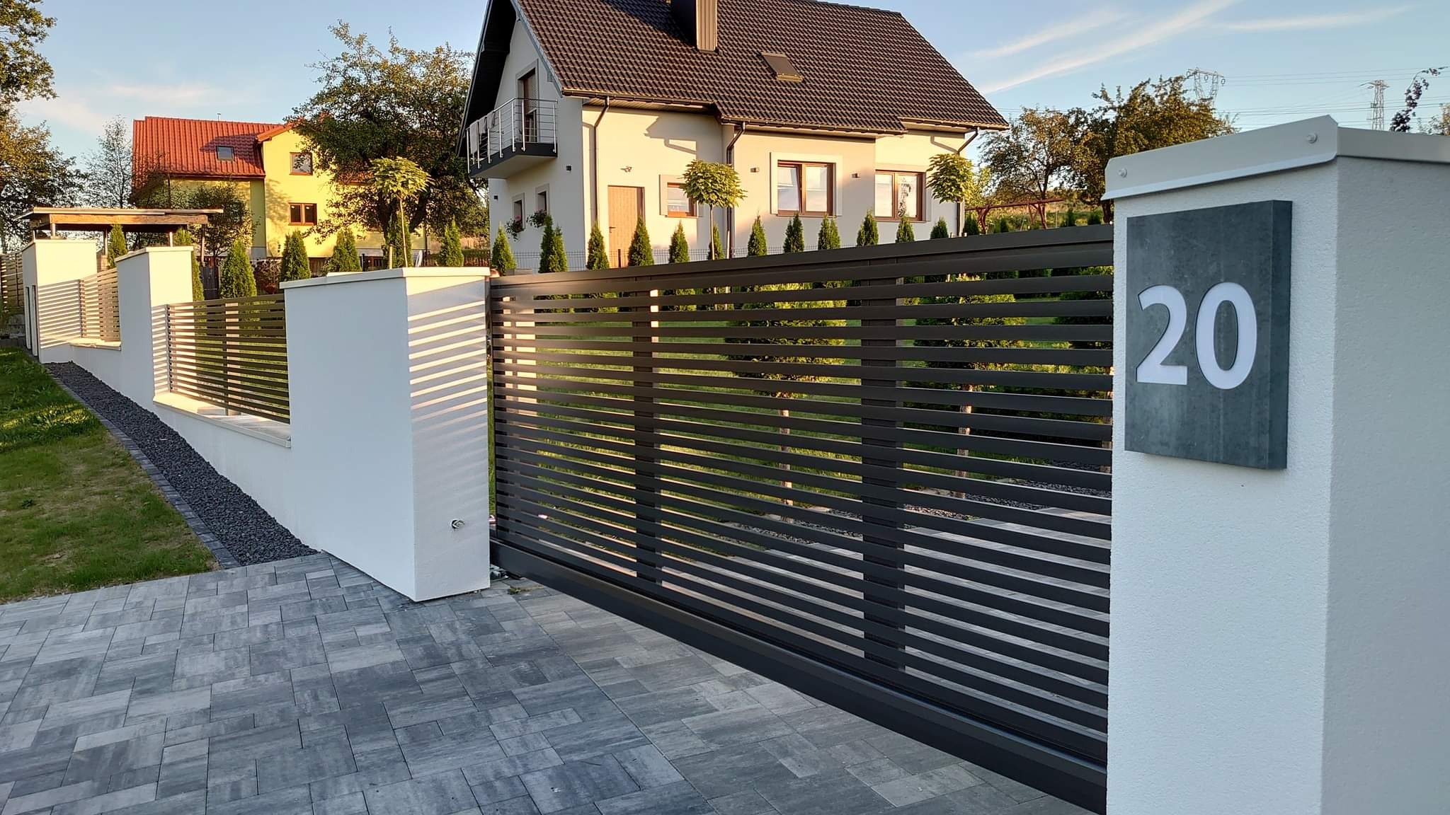 Nowoczesna, pozioma, przesuwna brama w kolorze antracytowym z numerem posesji '20' na białym słupku, na tle domu z ciemnym dachem i kostki brukowej.