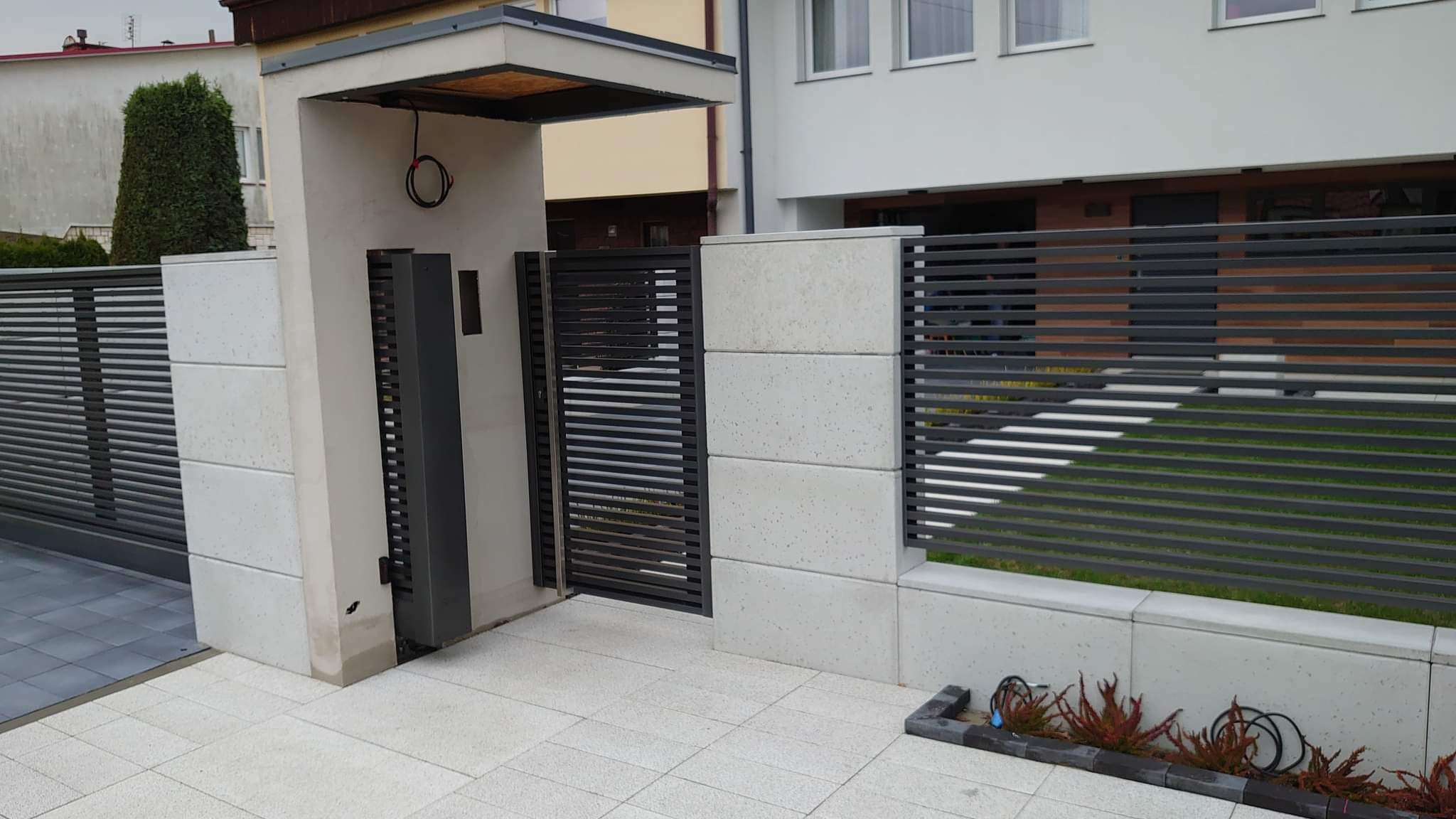 Nowoczesna, pozioma brama i ogrodzenie z metalowymi przęsłami w kolorze grafitowym, osadzone na betonowych słupkach i podmurówce w jasnym odcieniu szarości, z widocznym mechanizmem automatycznego...