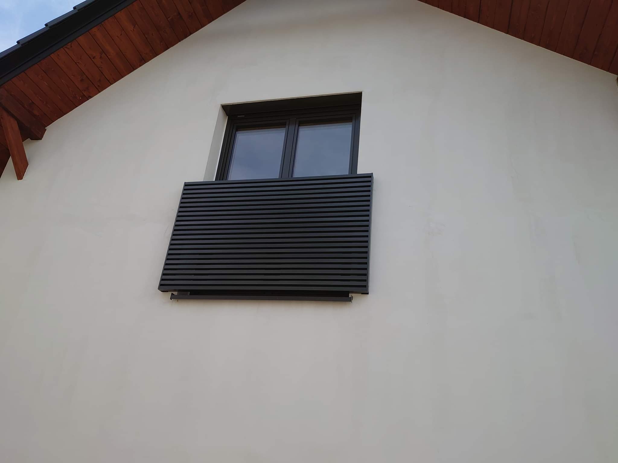Ciemnoszara, pozioma przesłona okienna z cienkich listew na elewacji jasnego domu z drewnianym okapem.