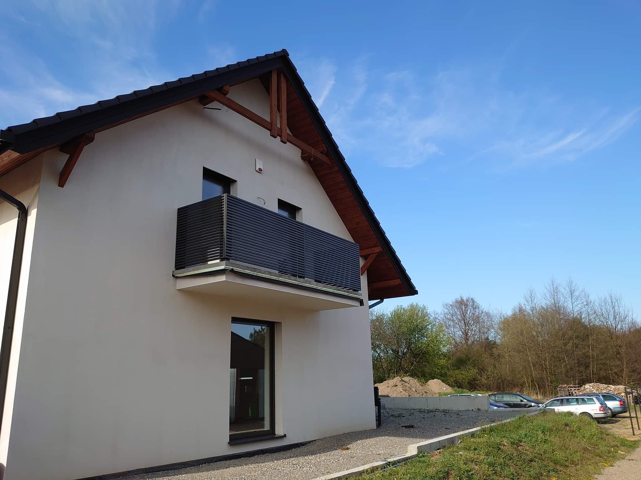 Nowoczesny dom jednorodzinny z balkonem zabezpieczonym poziomą, ciemną balustradą. Widoczny fragment elewacji, okno i zarys ogrodu z zaparkowanymi samochodami w tle.