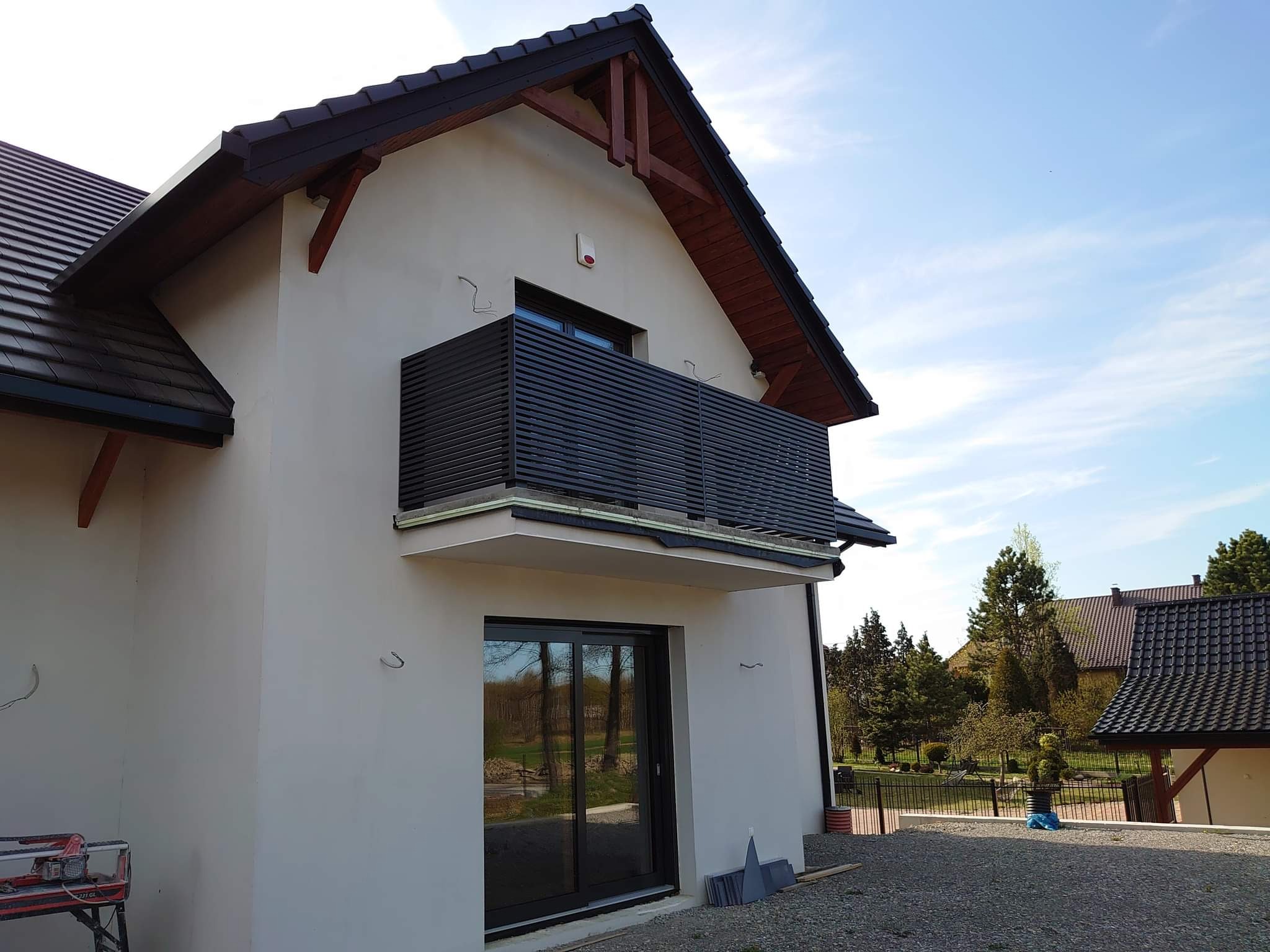 Nowoczesny balkon z poziomymi, ciemnymi listwami, widok z dołu na jasny budynek z ciemnym dachem i fragmentem podwórza z żwirowym podłożem oraz narzędzia budowlane w rogu.