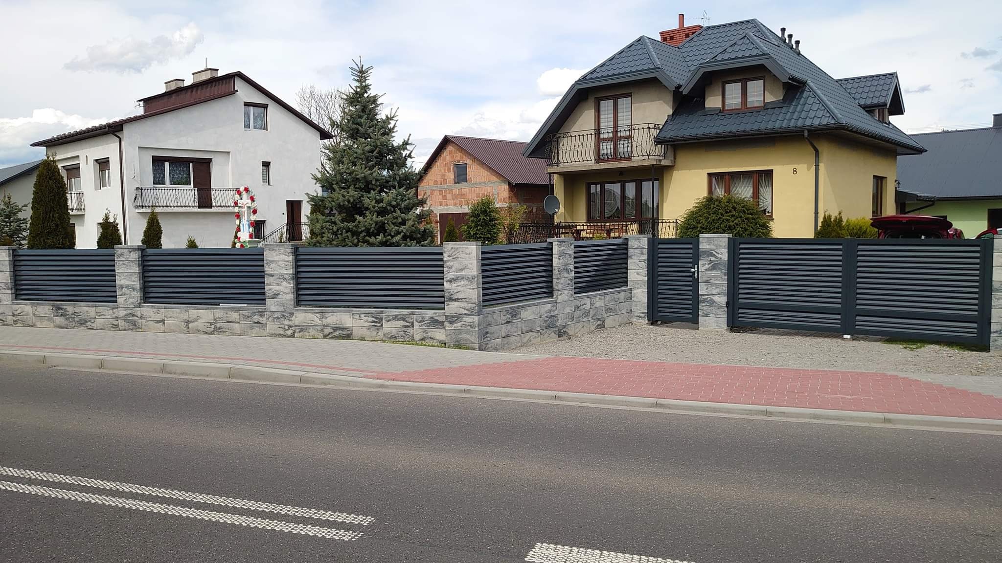 Nowoczesne, poziome, szare ogrodzenie z paneli metalowych z bramą wjazdową, osadzone na niskiej podmurówce z szarego kamienia łupanego, na tle domów jednorodzinnych.