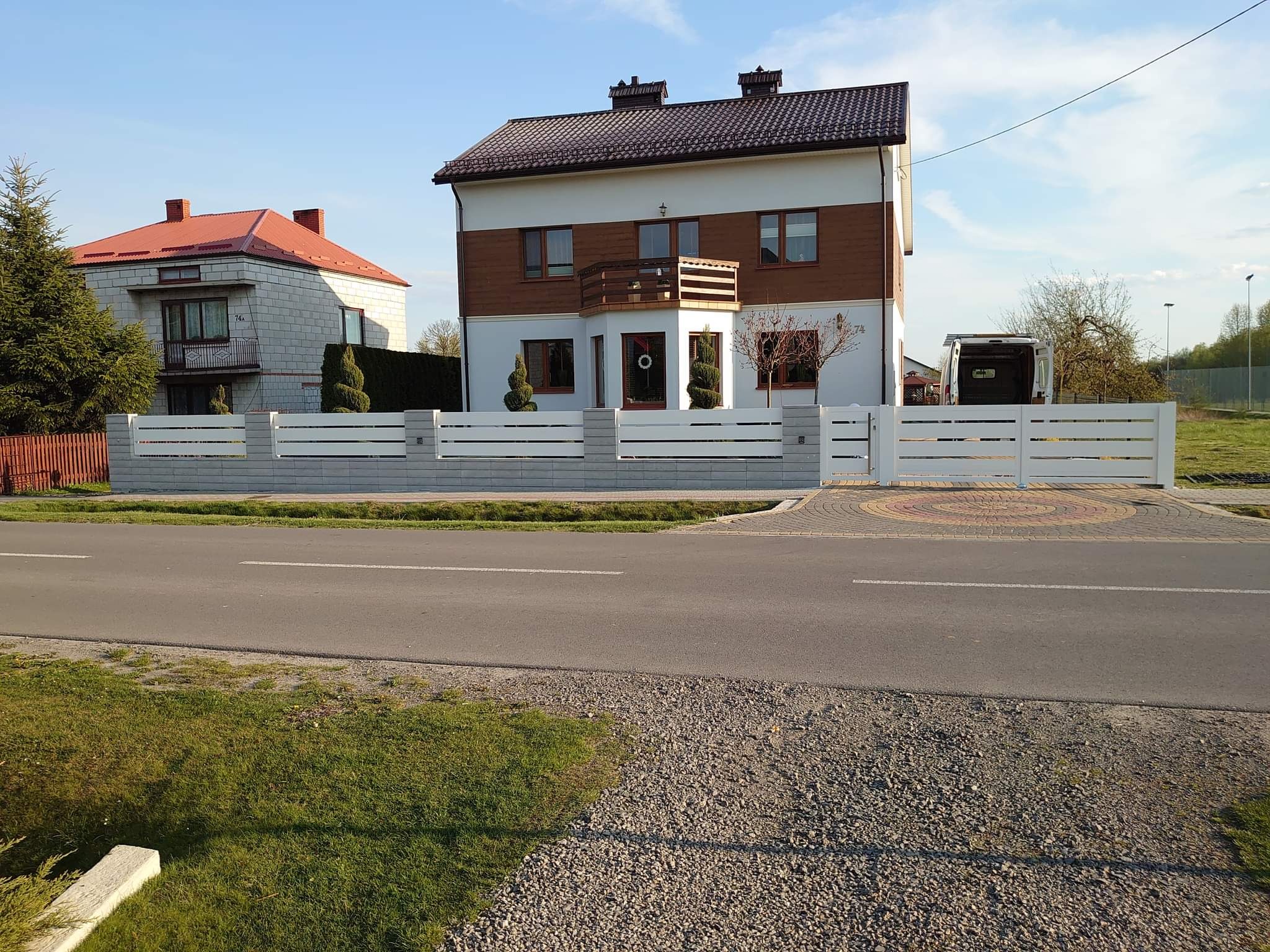 Nowoczesne ogrodzenie panelowe z białymi przęsłami i szarymi słupkami betonowymi przed dwupiętrowym domem z brązowymi elementami, widoczny samochód dostawczy w tle.