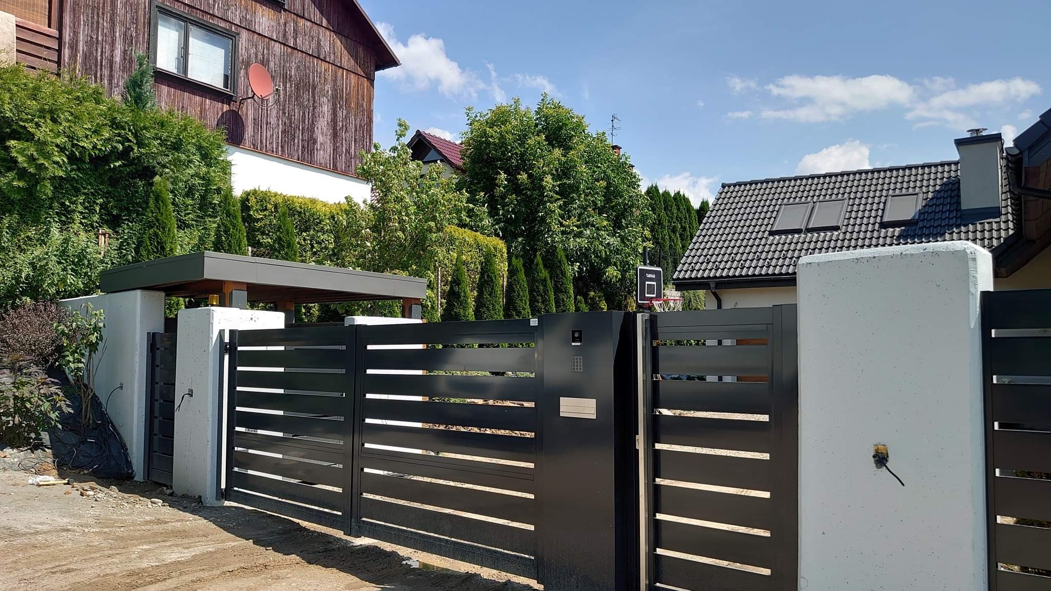 Nowoczesna, przesuwna brama metalowa w kolorze antracytowym z poziomymi panelami, zamontowana między betonowymi słupkami na tle zieleni i domów.