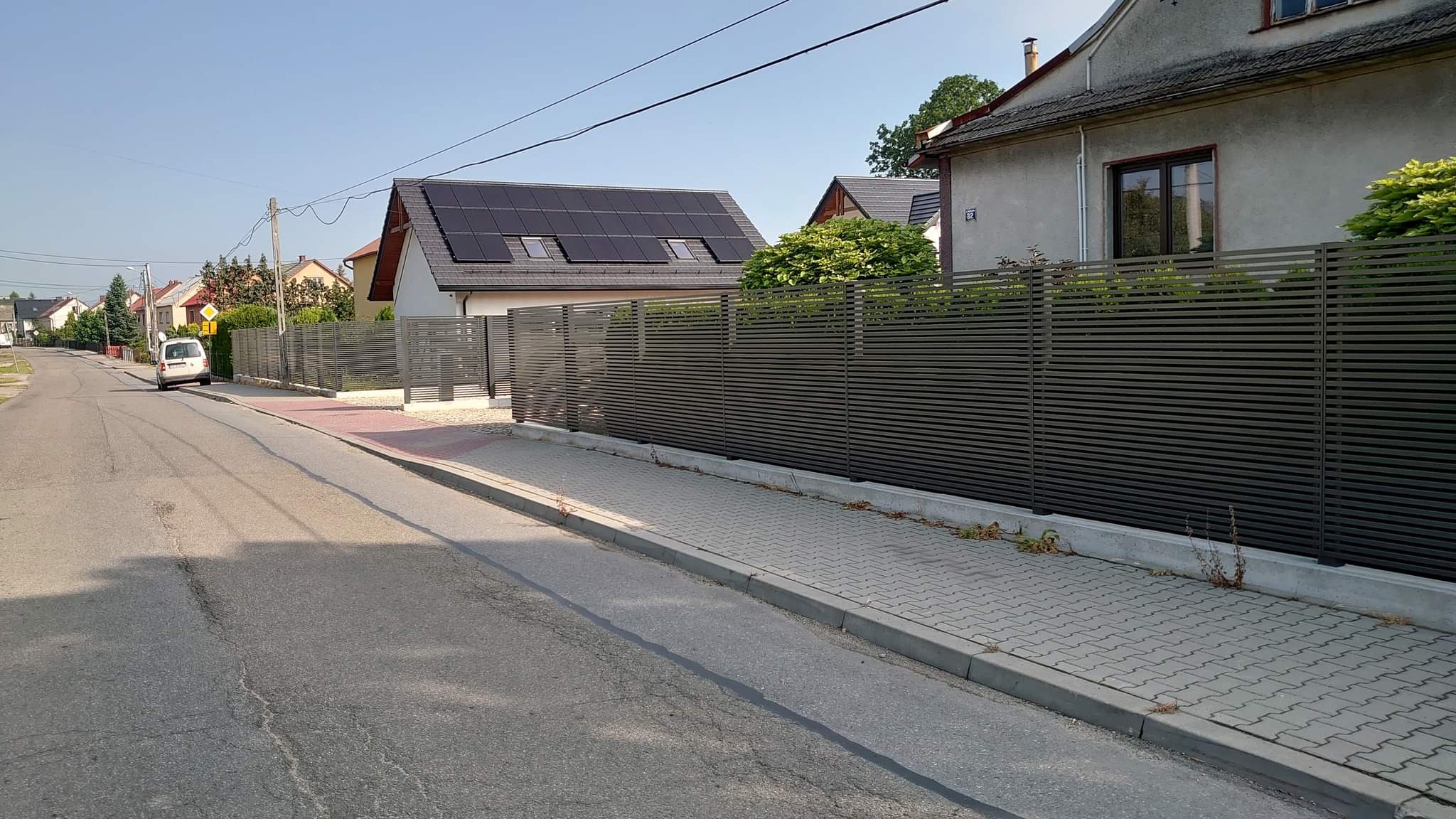 Długa, pozioma, szara osłona z listew tworząca ogrodzenie posesji z panelami słonecznymi na dachu domu, widok z ulicy.