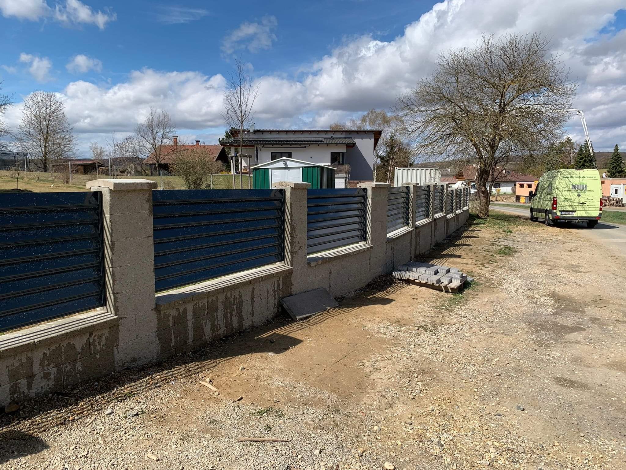 Nowy Targ: Fragment nowo wzniesionego, częściowo mokrego ogrodzenia z betonowymi słupkami i poziomymi, ciemnymi przęsłami. Obok leżą kostki brukowe, a w tle widać domy i częściowo zachmurzone niebo.