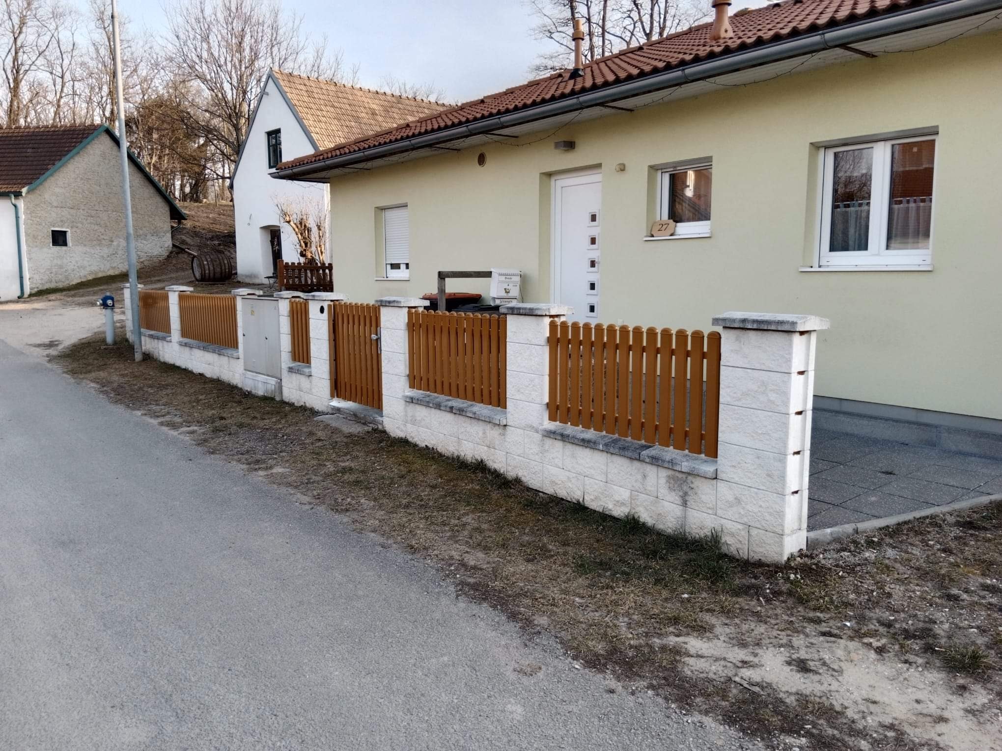 Nowoczesne ogrodzenie z drewnianymi przęsłami i kamiennymi słupkami przed domem z beżową elewacją i brązowym dachem, widok z poziomu ulicy.