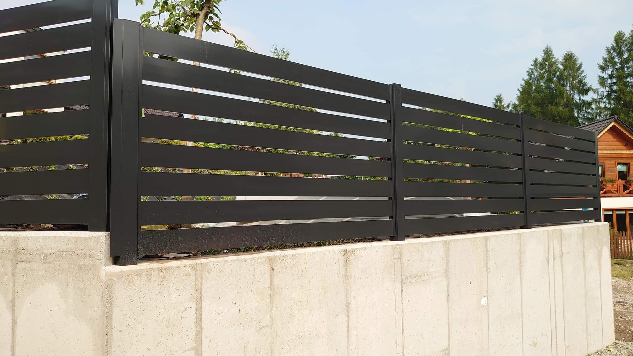 Nowoczesne, poziome, czarne ogrodzenie na betonowej podmurówce, z widokiem na drewniany dom i drzewa w tle.