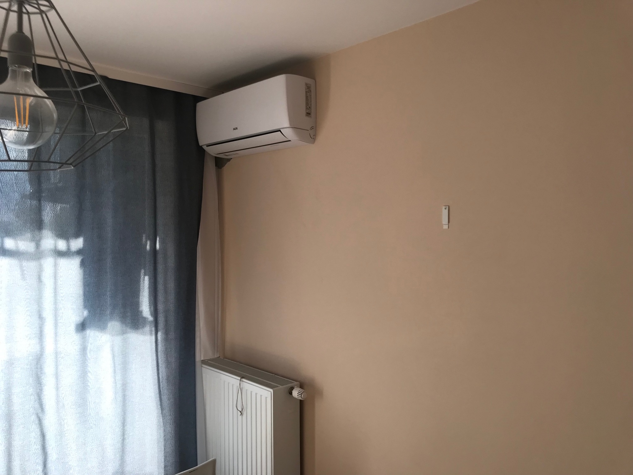 Biały klimatyzator zamontowany na beżowej ścianie w pokoju z szarą zasłoną, widoczny grzejnik i nowoczesna lampa wisząca