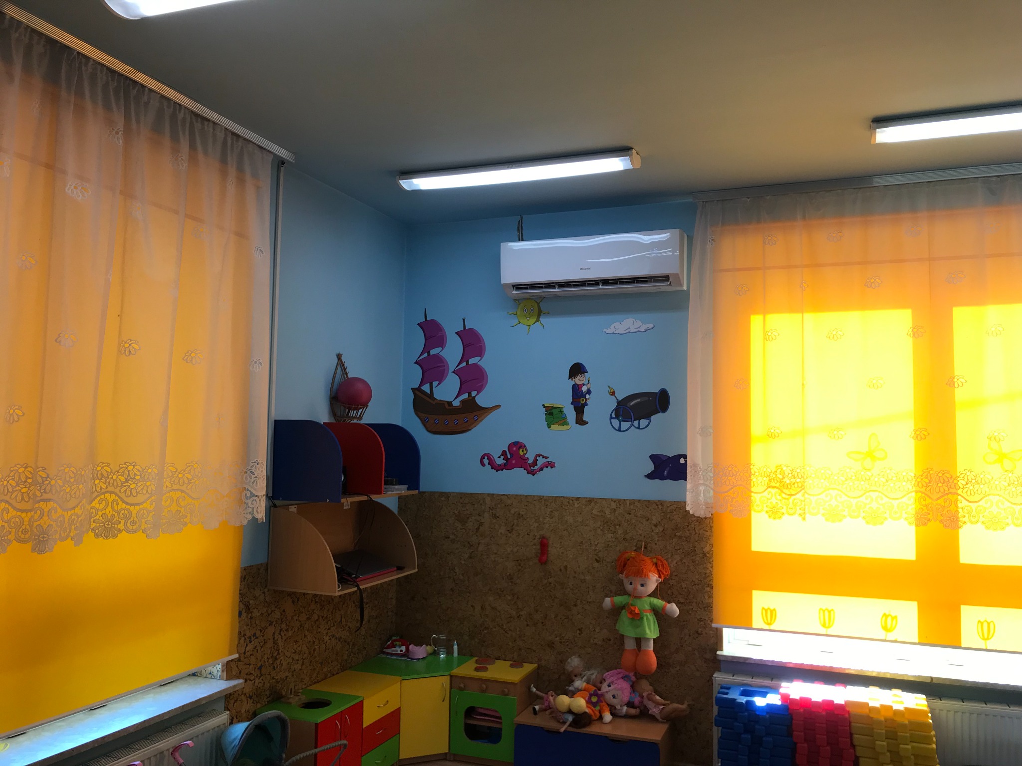 Klimatyzator ścienny zamontowany w pokoju dziecięcym z dekoracjami marynistycznymi, żółtymi zasłonami i zabawkami.