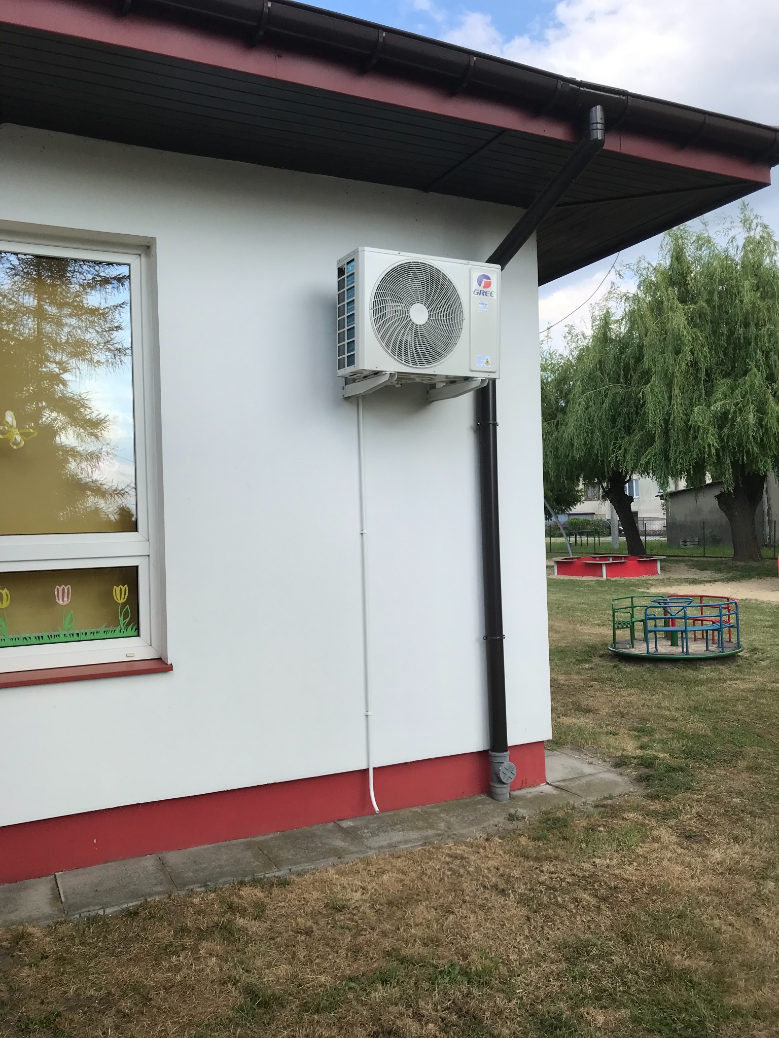 Biała jednostka zewnętrzna klimatyzatora marki Gree zamontowana na ścianie budynku z oknem ozdobionym rysunkami, widoczny rów spustowy i fragment placu zabaw z karuzelą.