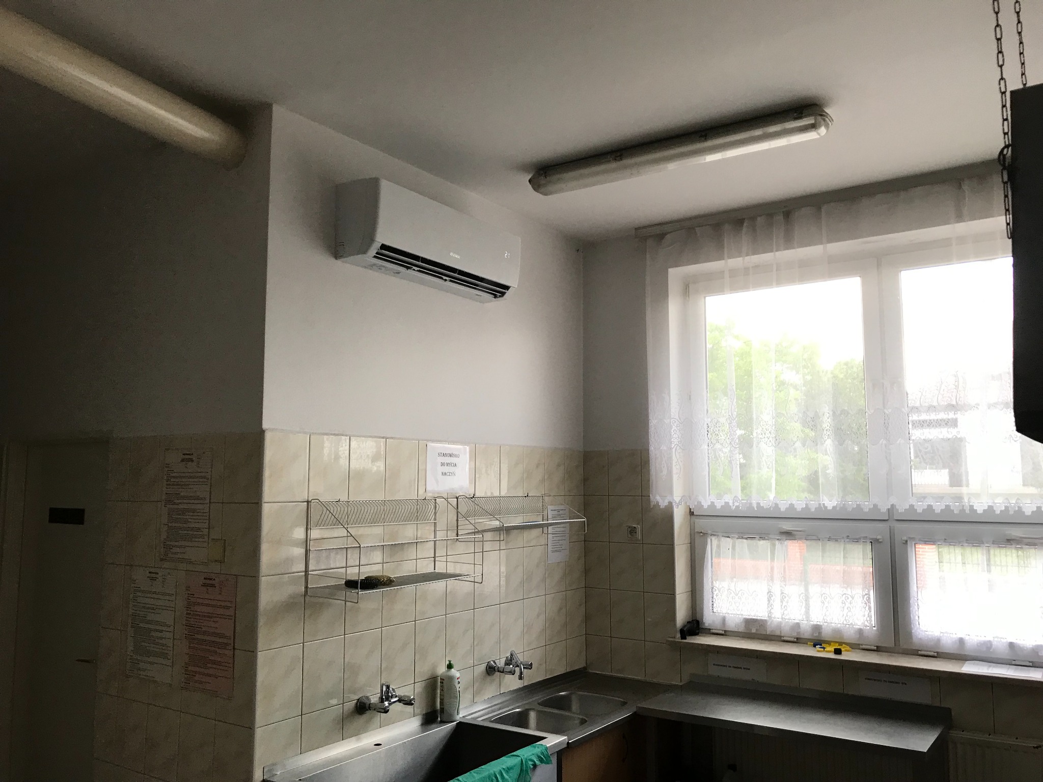 Biały klimatyzator ścienny zamontowany w pomieszczeniu z umywalkami i okładziną z płytek ceramicznych, widoczna suszarka na naczynia, okno z firanką.