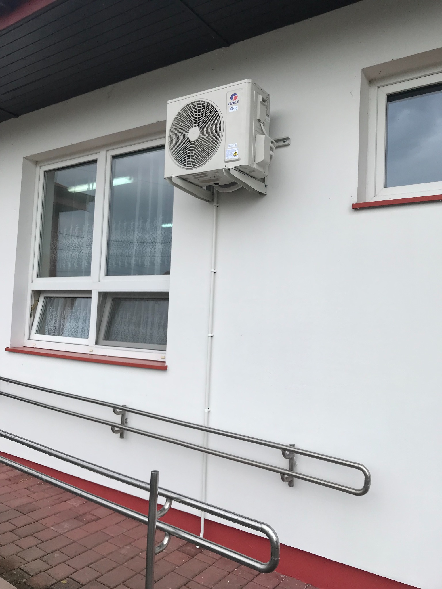 Biała jednostka zewnętrzna klimatyzacji marki Gree zamontowana na białej ścianie budynku obok okna z metalową barierką dla niepełnosprawnych.