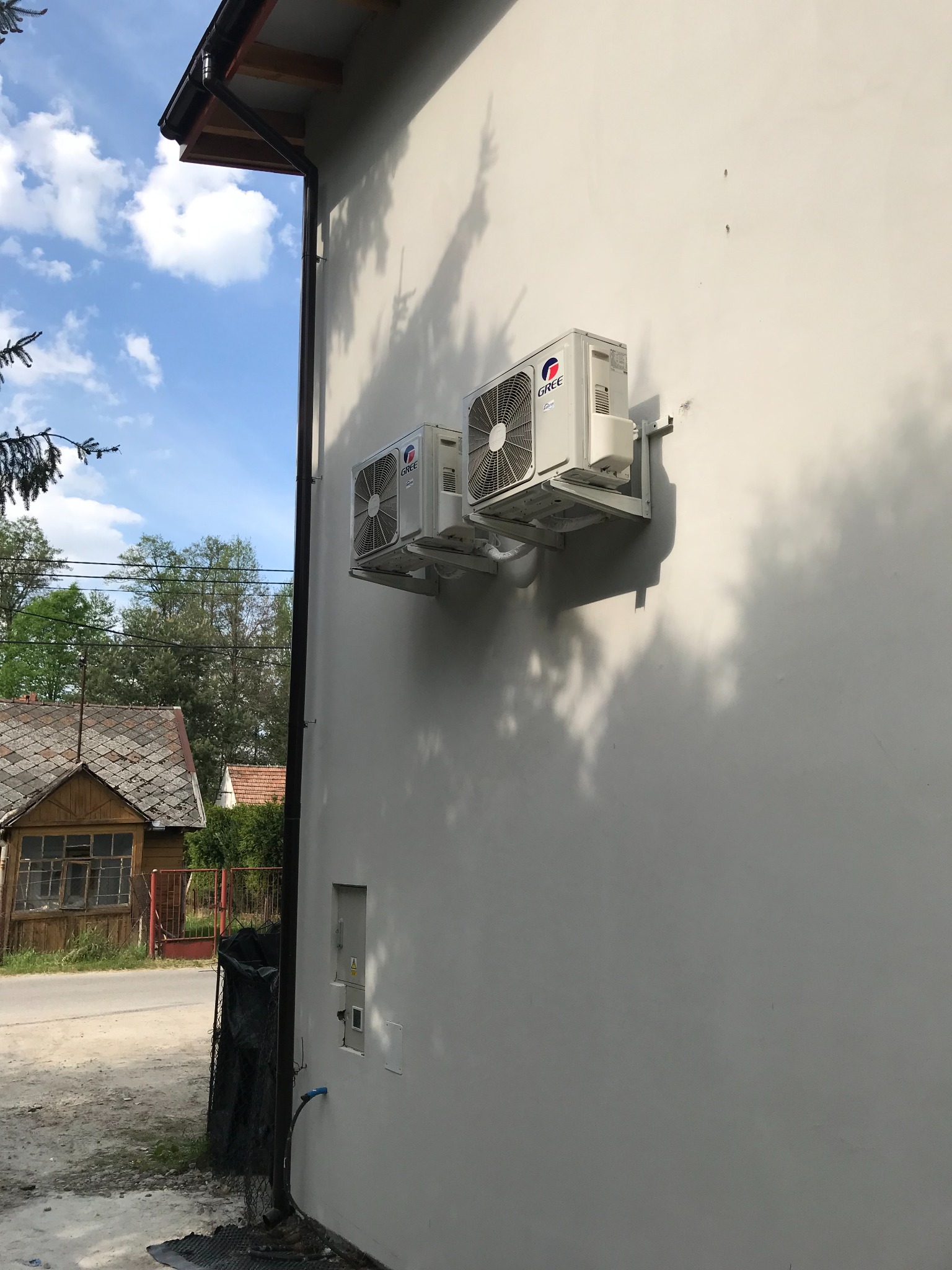 Dwa białe klimatyzatory marki Gree zamontowane na jasnej elewacji budynku mieszkalnego, widok z boku na tle błękitnego nieba i zabudowań wiejskich.
