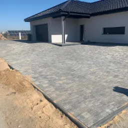 Nowa, szara kostka brukowa ułożona przed nowoczesnym domem z ciemną dachówką, widoczne przygotowane podłoże z piasku oraz stos drewna i panele słoneczne w tle.