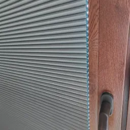 Szare plisowane osłony okienne zamontowane w drewnianej ramie okiennej z widoczną klamką.