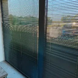 Ciemne, poziome lamele w oknie z czarną ramą, widok z wnętrza pomieszczenia na zewnątrz, delikatne odbicie światła na powierzchni, szara, kamienna podłoga w tle.