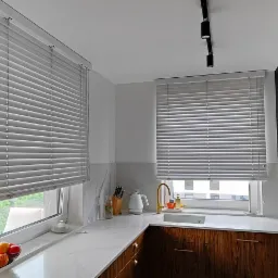 Nowoczesna kuchnia z białymi poziomymi osłonami okiennymi, złotym okapem i armaturą, ciemnymi szafkami z podłużnymi uchwytami oraz marmurowym blatem i backsplashiem.