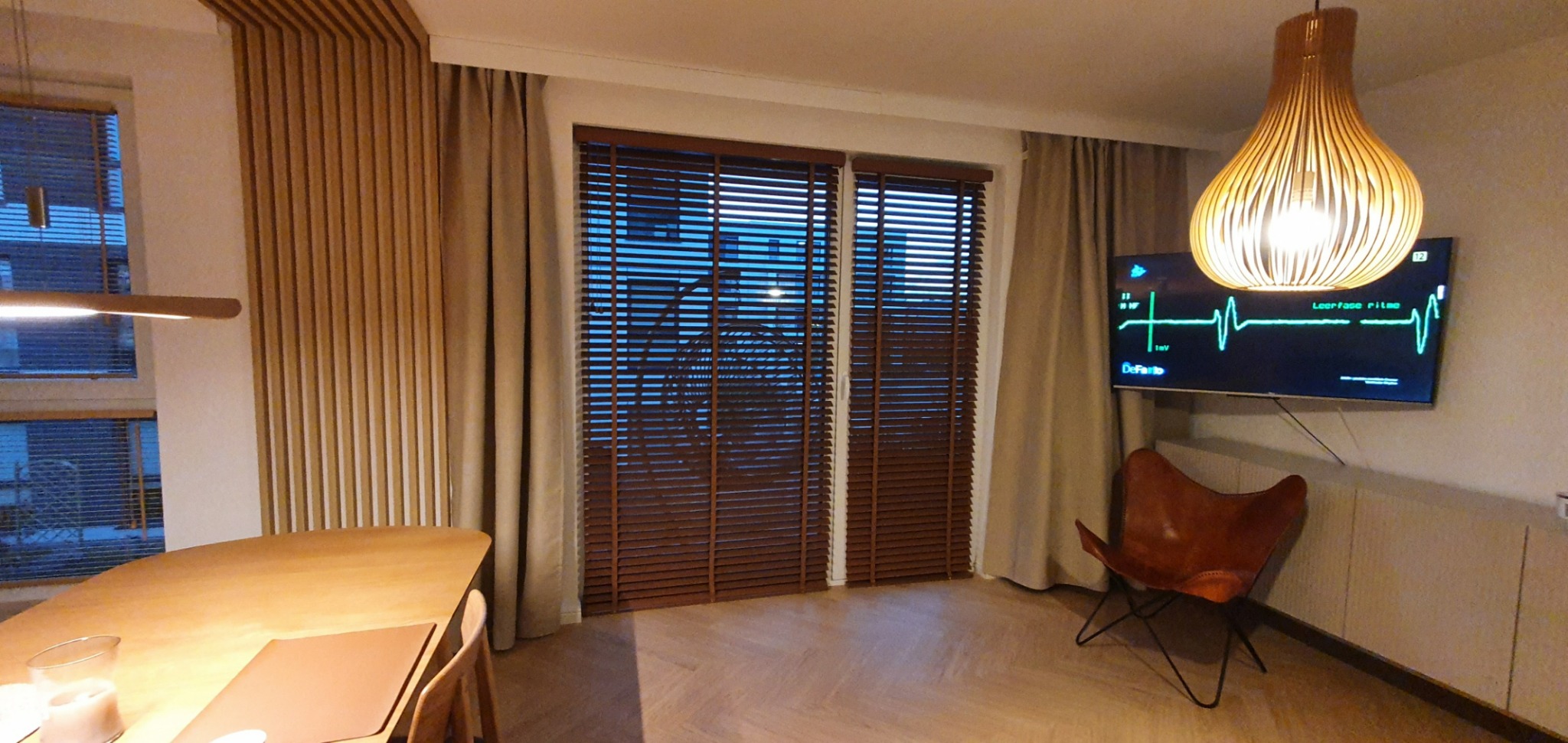 Jasne wnętrze z drewnianym stołem, designerską lampą i oknami z ciemnymi poziomymi przesłonami; na ścianie telewizor z wyświetlanym wykresem EKG.