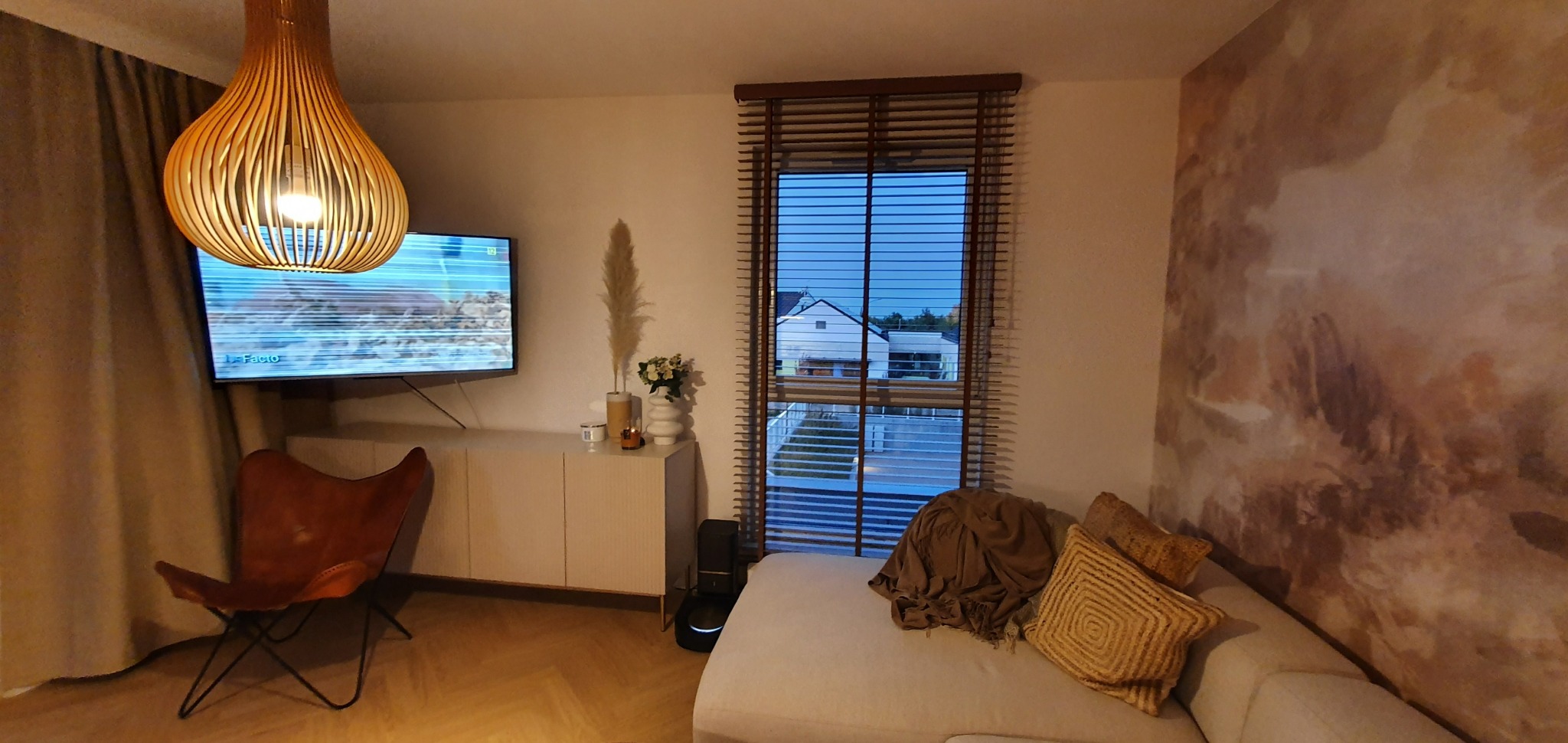 Salon z drewnianymi żaluzjami poziomymi w oknie, przez które widać dom i ogród; ciepłe oświetlenie, dekoracyjna lampa wisząca, telewizor, kanapa z poduszkami i fotel motylkowy.