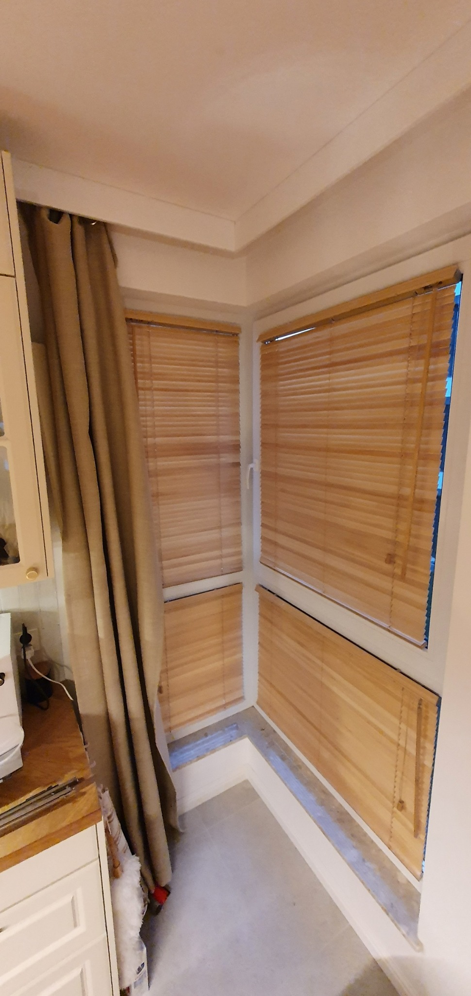 Fragment wnętrza z oknami wyposażonymi w poziome przesłony okienne z drewnopodobnego materiału, częściowo zasłonięte beżową zasłoną, widok z bliska.