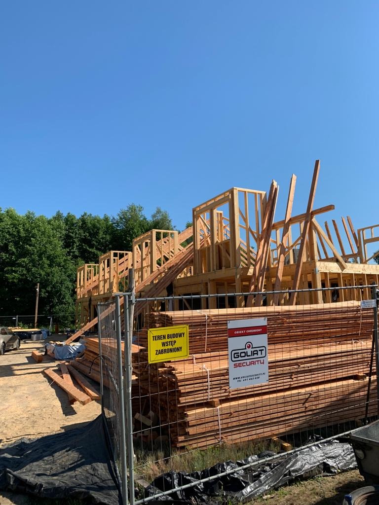 Wznoszona konstrukcja domu z drewna, widok zza ogrodzenia budowy z tabliczką ostrzegawczą i stosem desek.