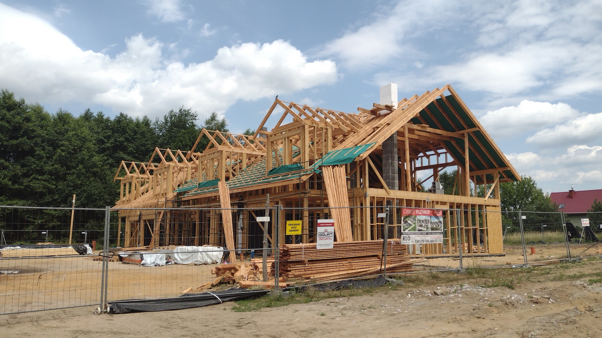 Konstrukcja domu szkieletowego w trakcie budowy, widoczne drewniane elementy szkieletu, komin z szarej cegły, zabezpieczona siatką budowa, stos desek i tablice informacyjne.