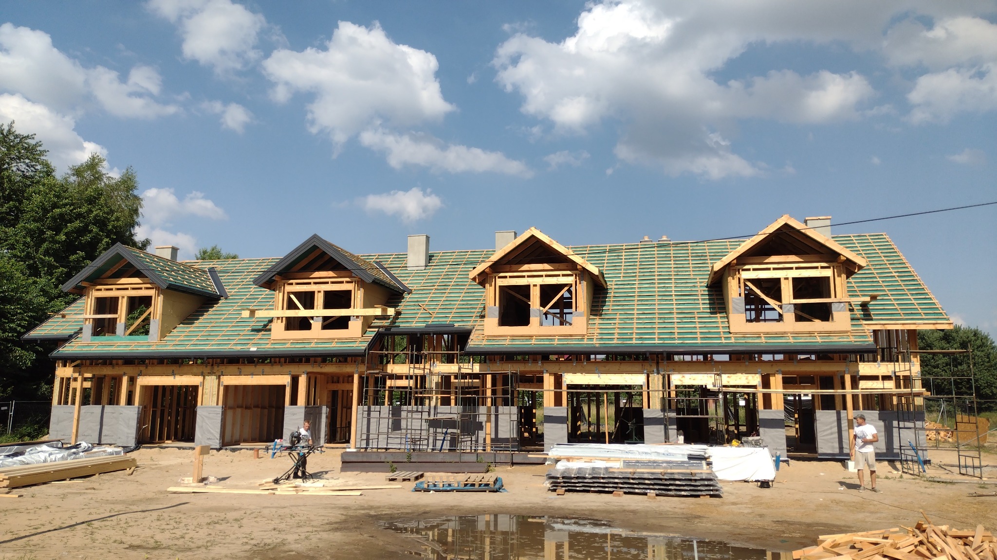 Budowa domu z drewnianą konstrukcją szkieletową, widoczne elementy dachu i ścian zewnętrznych, pracownik przy pile ukośnicy, materiały budowlane na placu budowy.