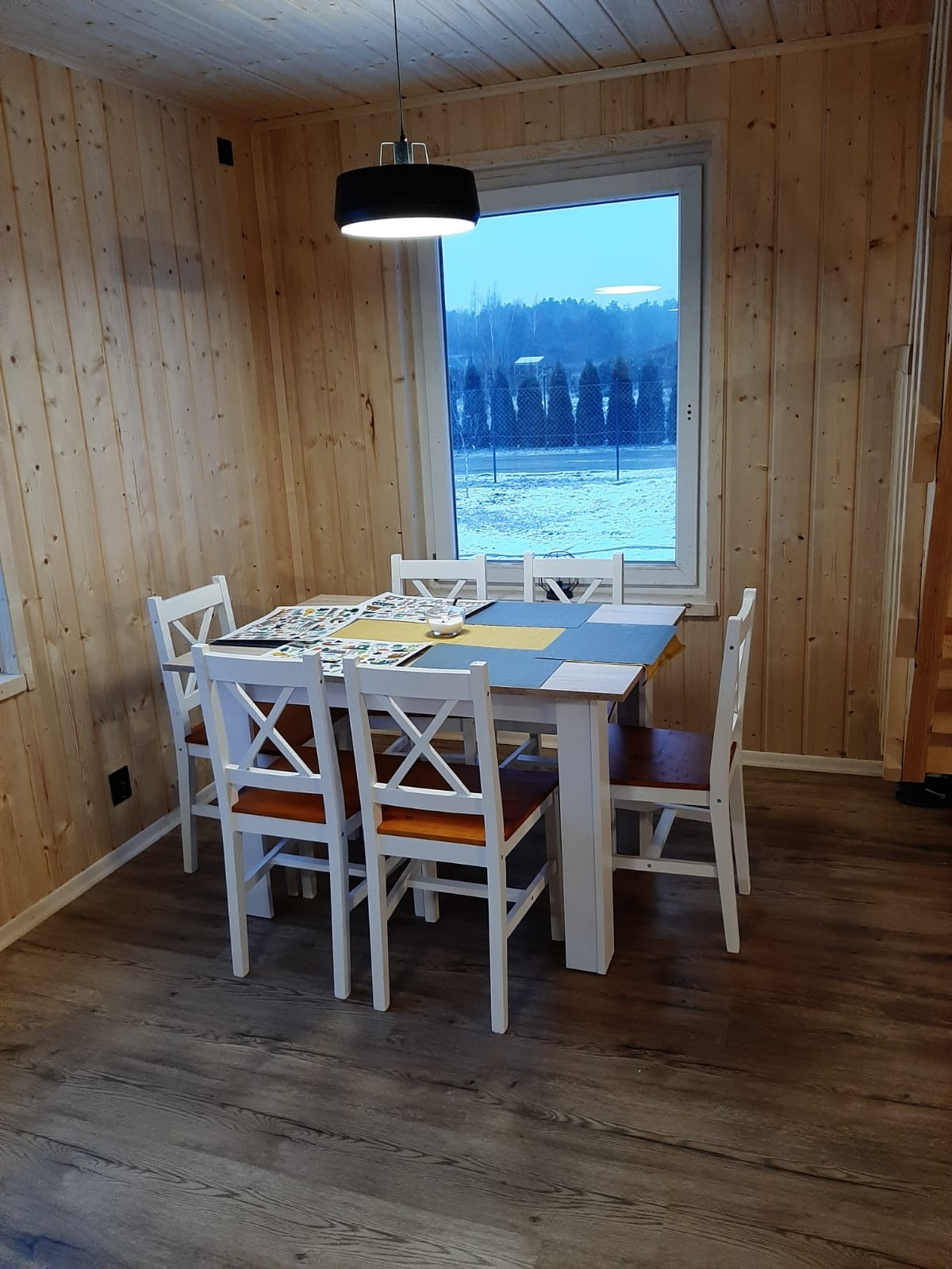 Wnętrze z drewnianą boazerią: Stół z białymi krzesłami, nakryty kolorową serwetą, widok na zimowy krajobraz za oknem.