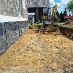 Przygotowanie terenu pod fundamenty domu: wykop z żółtym piaskiem, zabezpieczona ściana budynku z izolacją, mała koparka w tle.