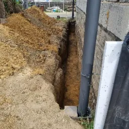 Wąski wykop wzdłuż ściany budynku z widoczną rurą spustową i stertą ziemi, przygotowany prawdopodobnie pod izolację fundamentów.