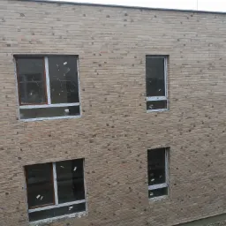 Nowo wymurowana ściana budynku z cegły klinkierowej w odcieniach beżu i brązu, z zamontowanymi oknami o r&oacute;żnej wielkości, zabezpieczonymi folią. Widoczne nier&oacute;wności w fugach i zaprawa murarska.