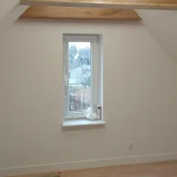 Poddasze po remoncie: jasne ściany, drewniane belki stropowe z oświetleniem, okno z widokiem na zewnątrz, nowa podłoga w panelach imitujących drewno, zabezpieczona folią malarską.