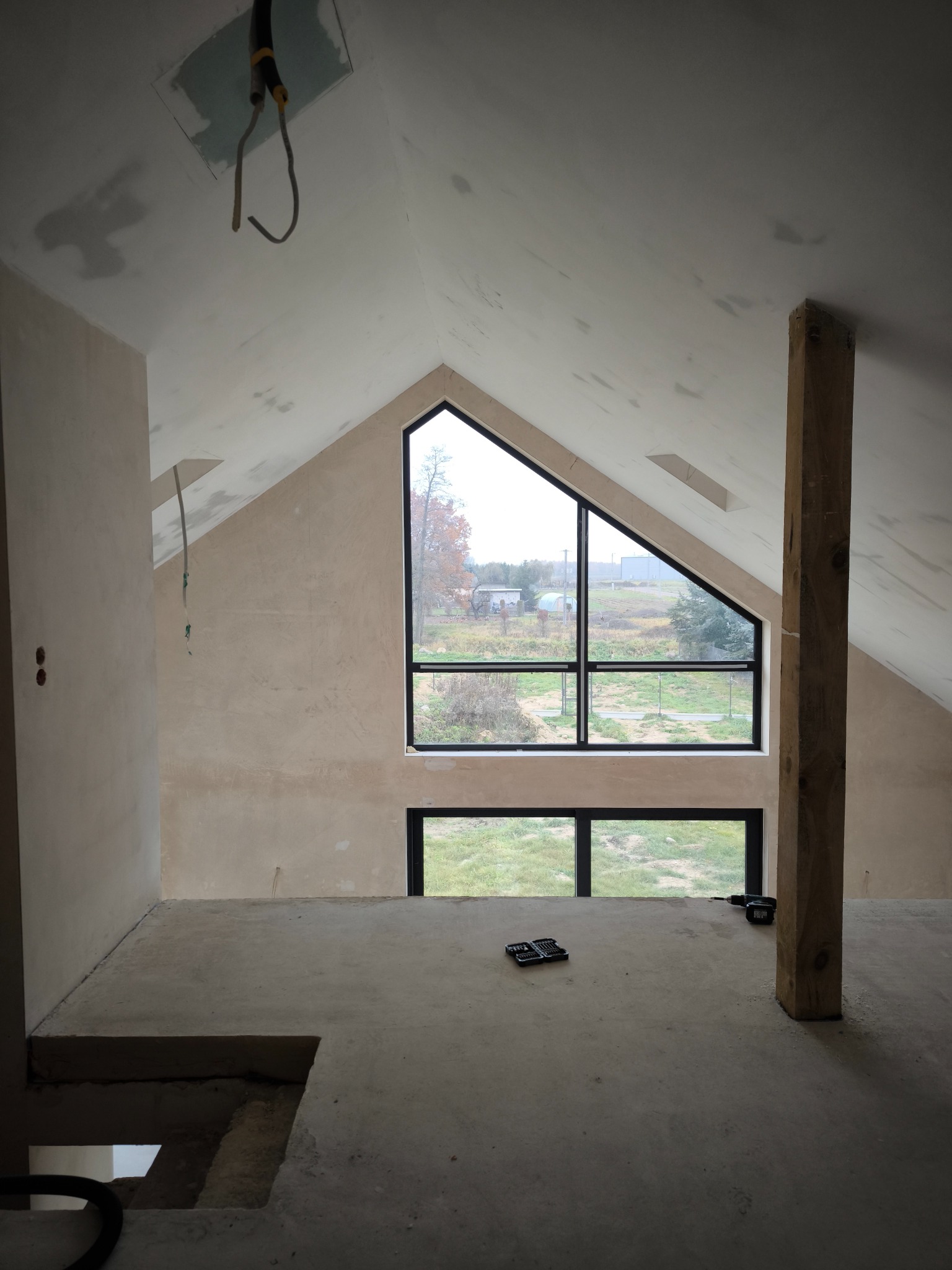 Poddasze w trakcie wykończenia: ściany po gładzi, surowa podłoga, okno trójkątne z czarną ramą, widoczny słup drewniany i narzędzia na podłodze.