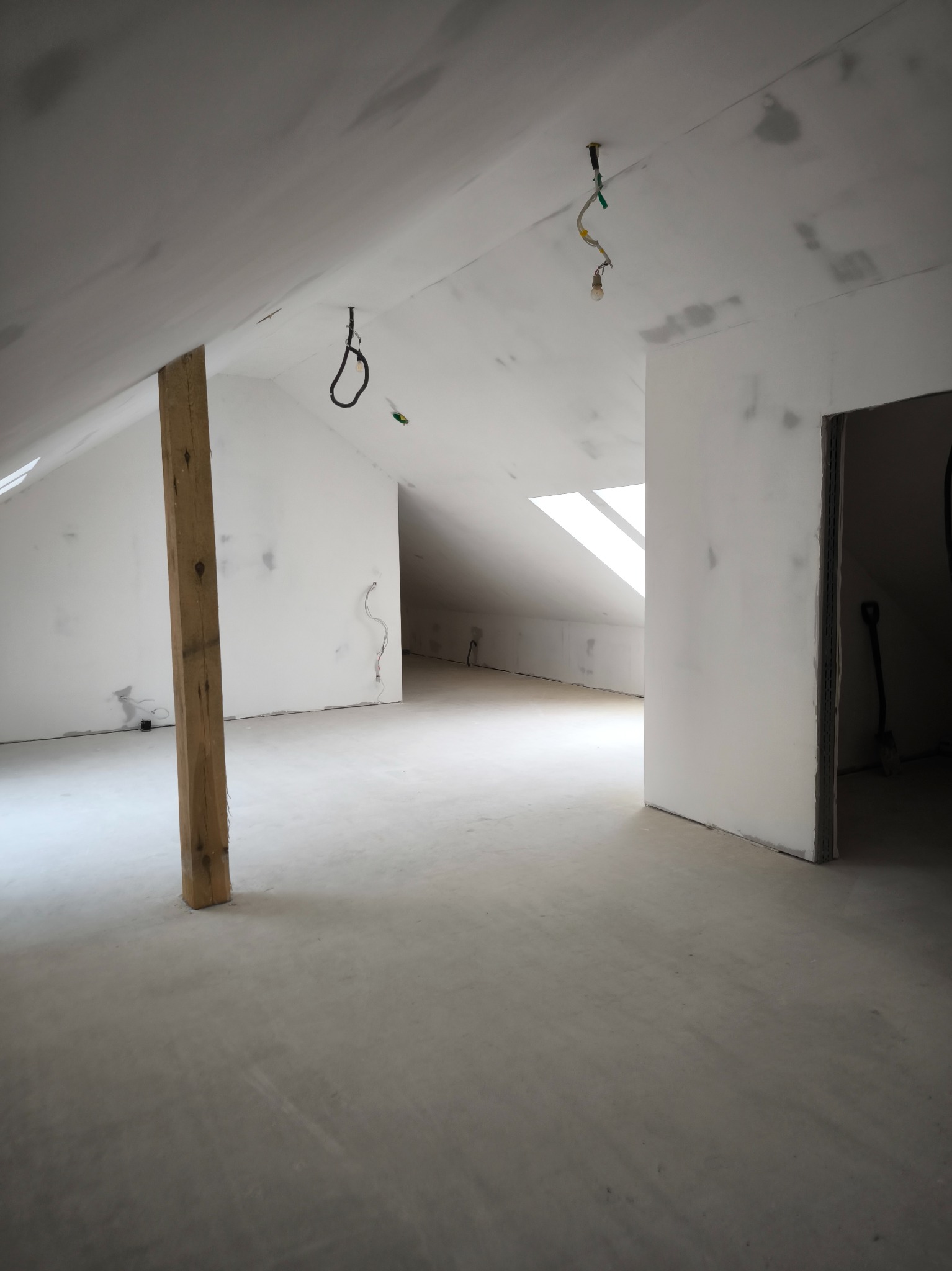 Poddasze w trakcie prac wykończeniowych, widoczne ślady po gruntowaniu ścian i sufitu, drewniany słup konstrukcyjny, okno dachowe i wiszące przewody elektryczne.