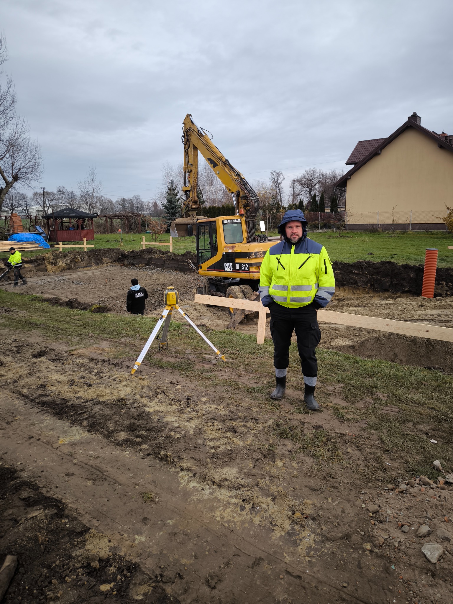 Pracownik w odblaskowej kurtce stoi na placu budowy z wykopem pod fundamenty, w tle koparka CAT M 312 i niwelator laserowy na statywie.