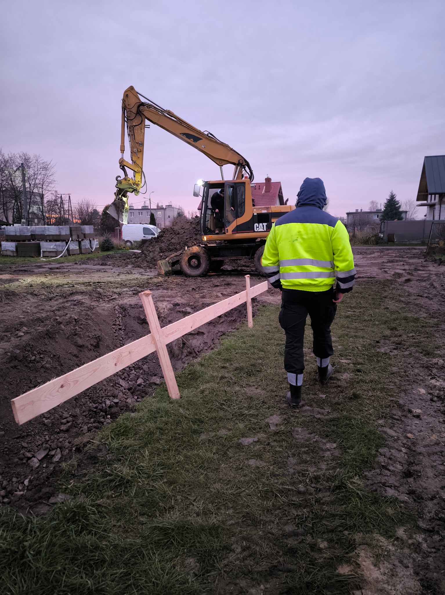 Żółta koparka CAT w trakcie prac ziemnych przy wykopie, widoczny pracownik w odblaskowej kurtce i drewniane szalunki