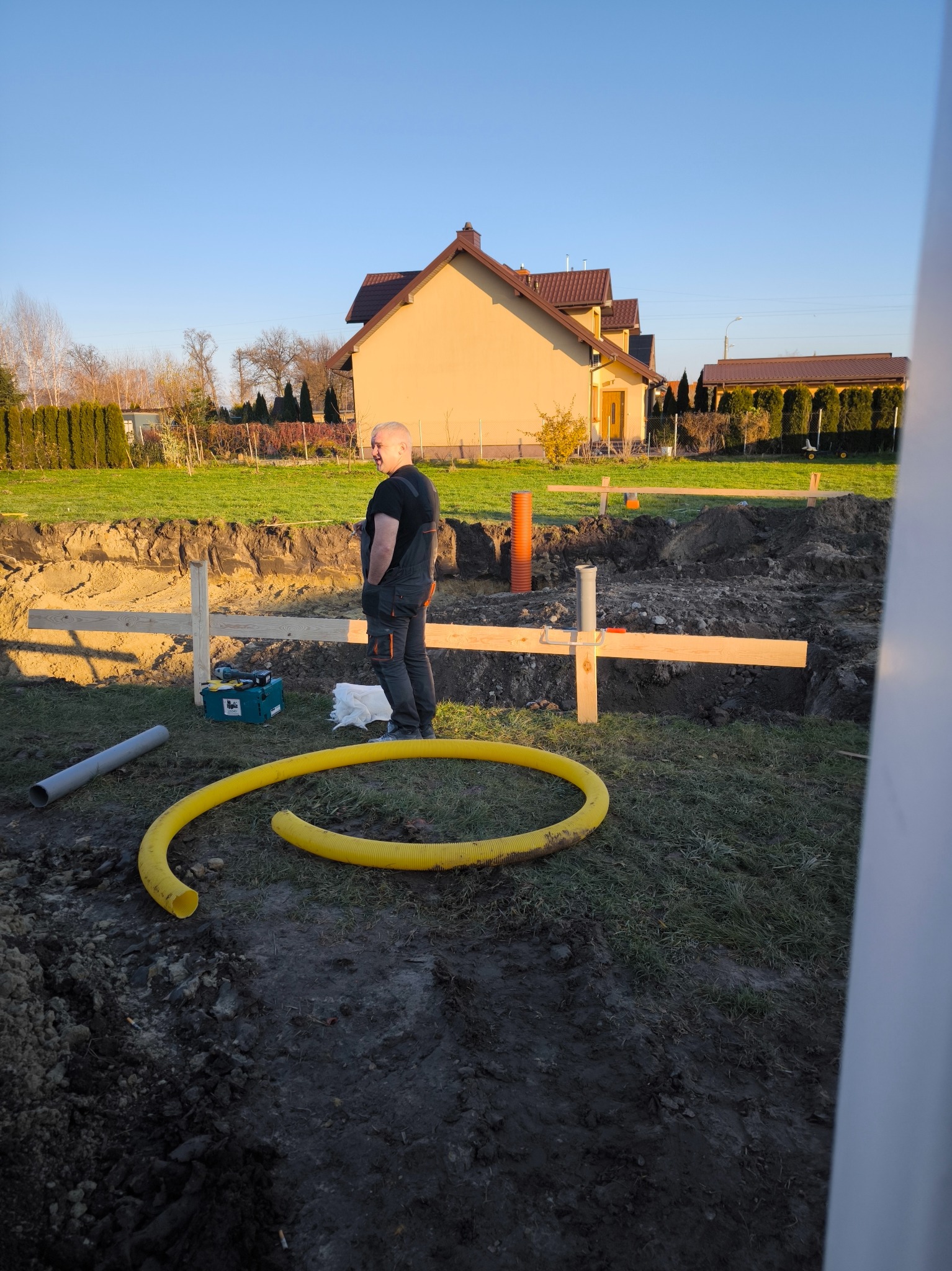 Fundament pod budynek jednorodzinny w trakcie budowy, widoczne drewniane szalunki, rury kanalizacyjne i pracownik w kombinezonie roboczym na tle domu w słoneczny dzień.