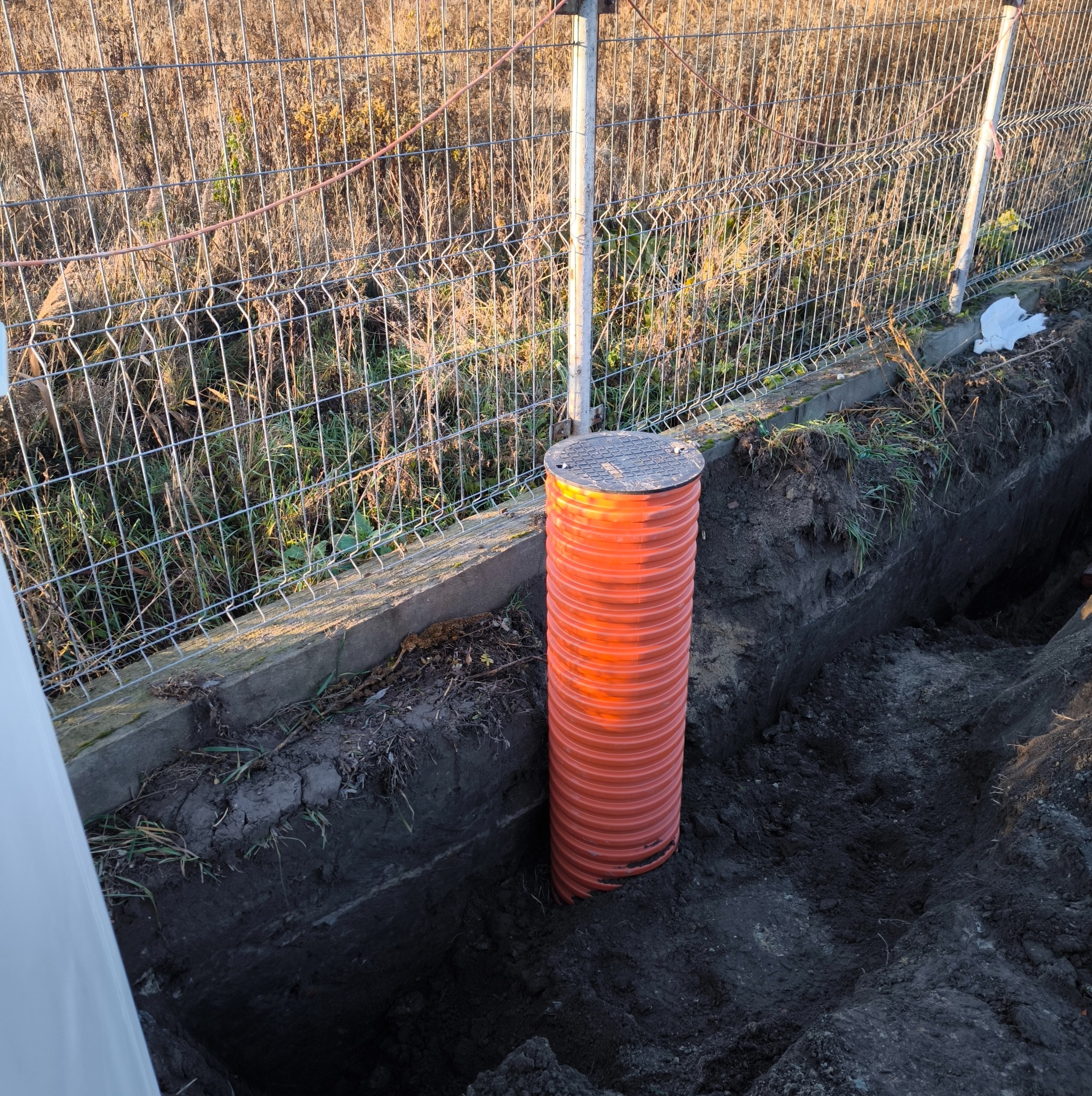 Pomarańczowa rura kanalizacyjna z szarą pokrywą wystaje z wykopu, widoczna siatka ogrodzeniowa w tle.