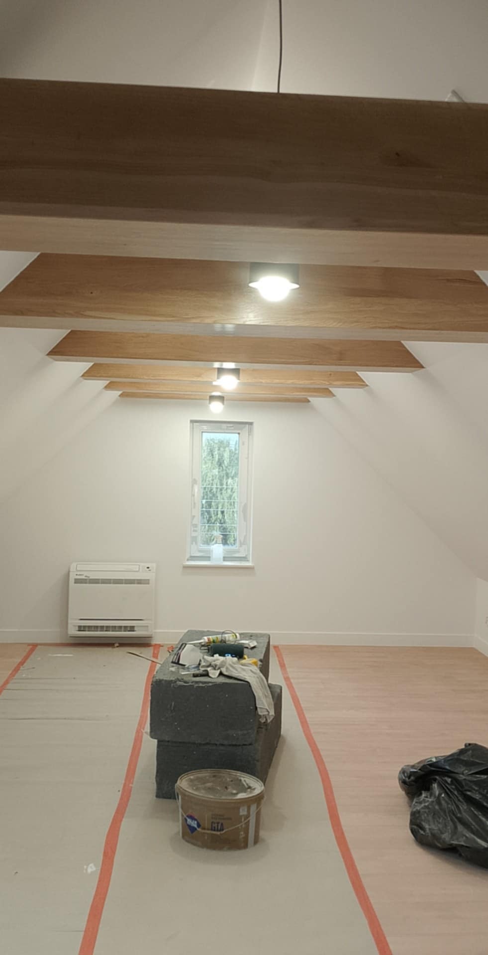 Poddasze z drewnianymi belkami stropowymi i oświetleniem punktowym, przygotowane do prac wykończeniowych, z zabezpieczoną podłogą i blokami styropianowymi jako podstawą roboczą.