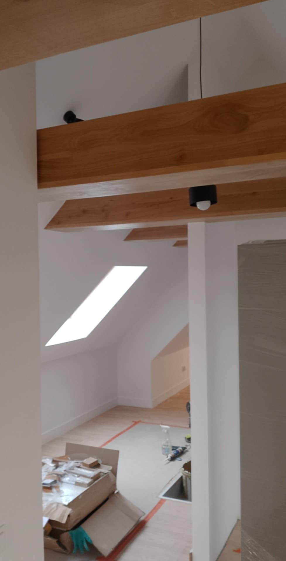 Poddasze z drewnianymi belkami stropowymi, oknem dachowym i widocznymi pracami wykończeniowymi w trakcie realizacji, w tym kartonami i narzędziami na podłodze.