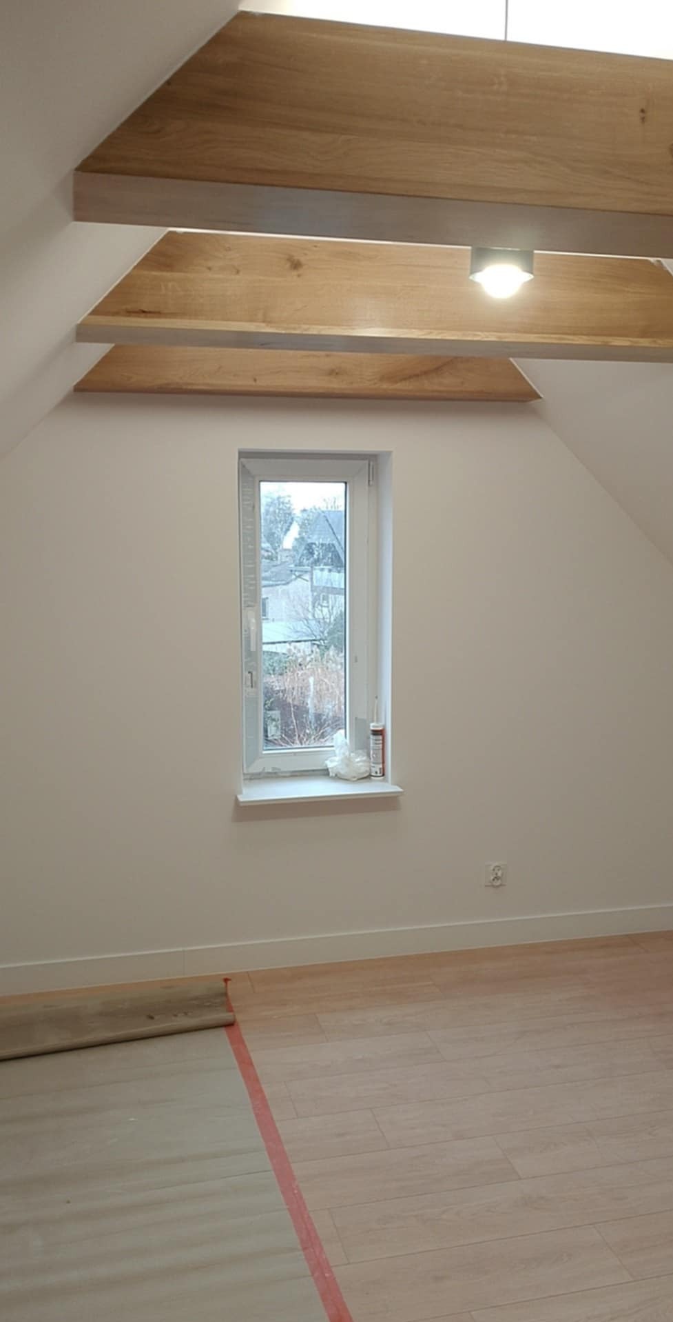 Poddasze po remoncie: jasne ściany, drewniane belki stropowe z oświetleniem, okno z widokiem na zewnątrz, nowa podłoga w panelach imitujących drewno, zabezpieczona folią malarską.