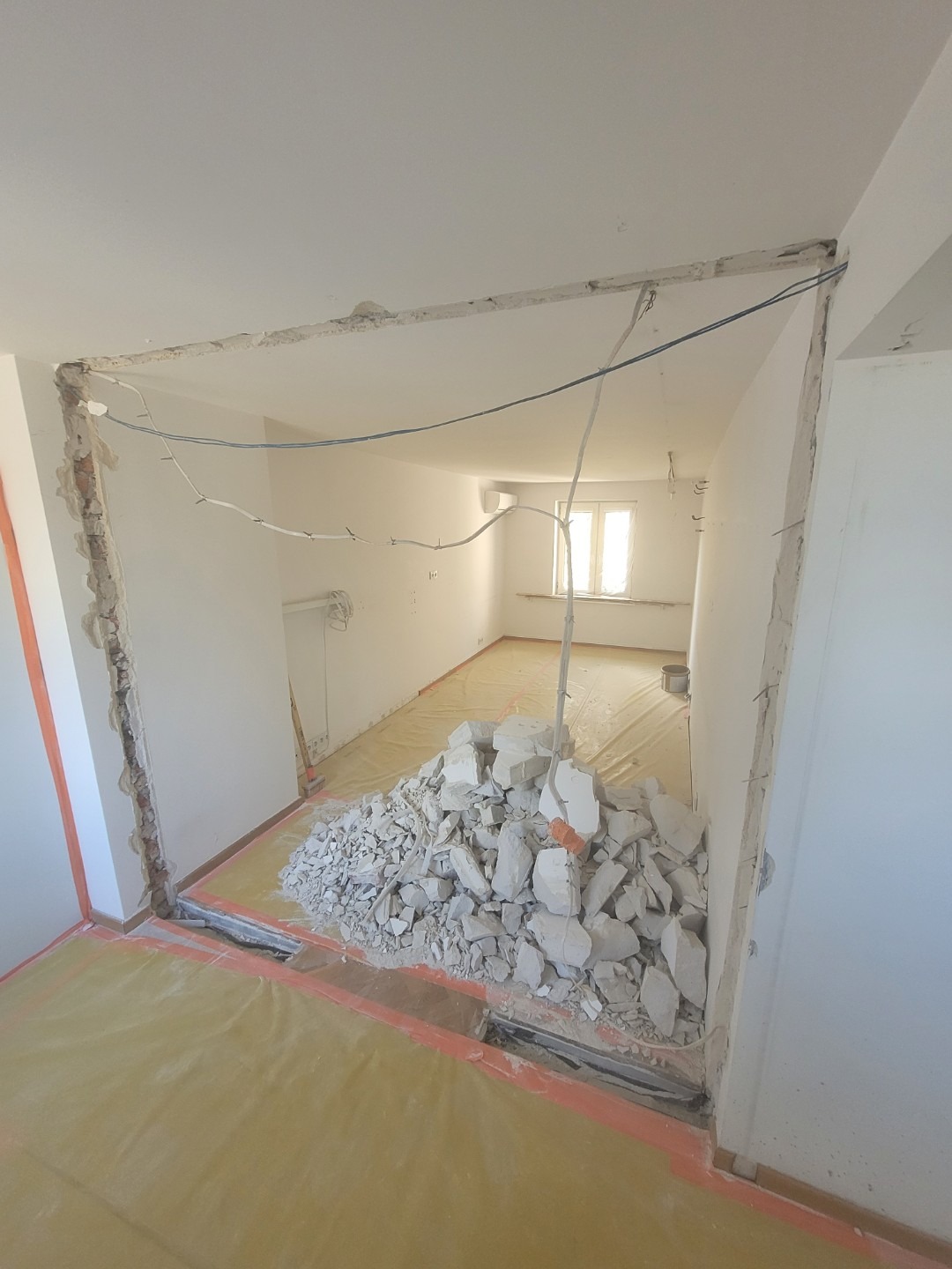 Demontaż ściany działowej w mieszkaniu, widoczne gruzy i zabezpieczona folią podłoga. Prace remontowe w trakcie realizacji. Widoczne przewody elektryczne.
