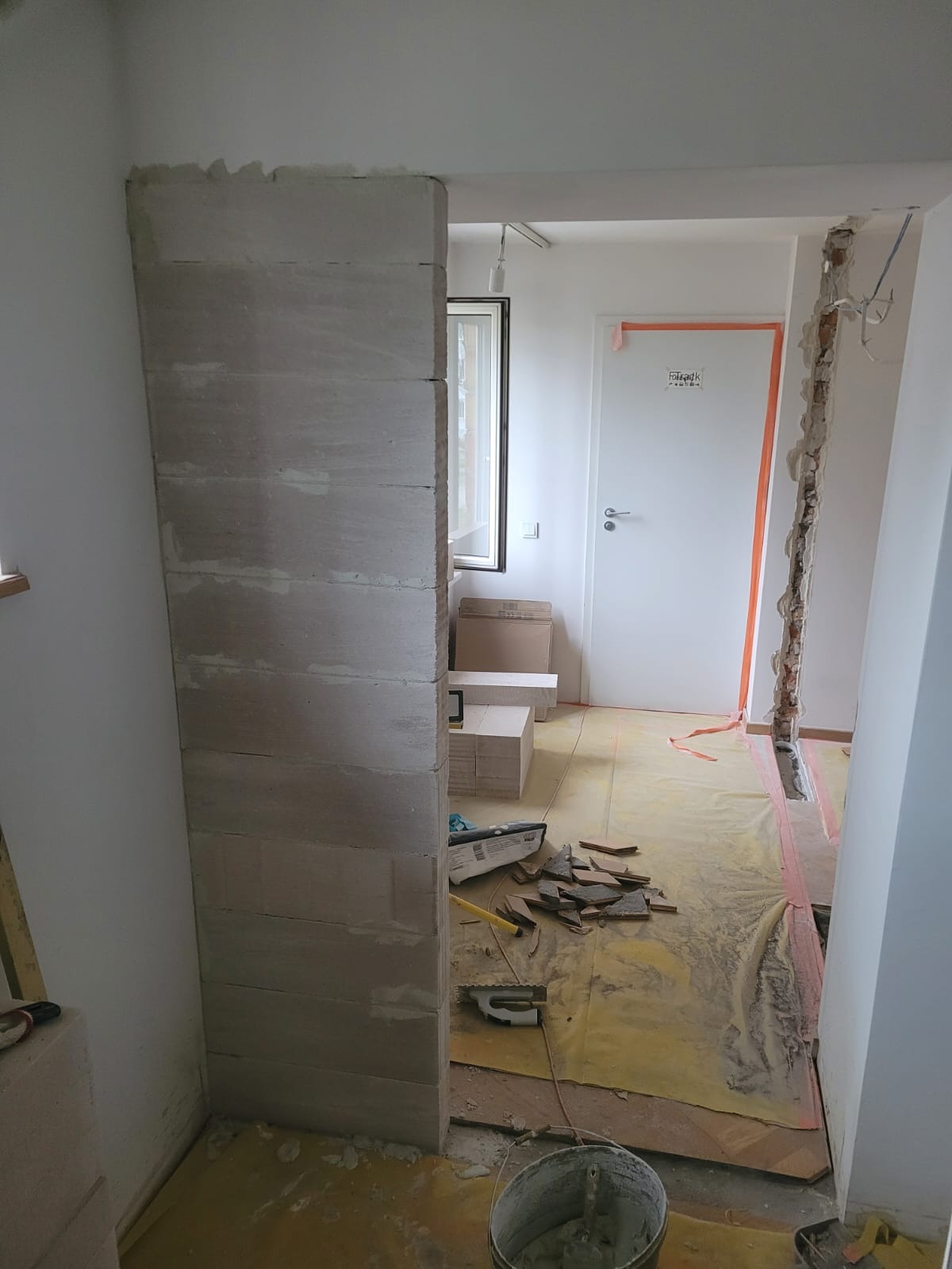 Murowana ścianka działowa z betonu komórkowego na żółtej folii ochronnej. W tle drzwi i instalacja w ścianie. Widoczne narzędzia i wiadro. Prace wykończeniowe w trakcie.