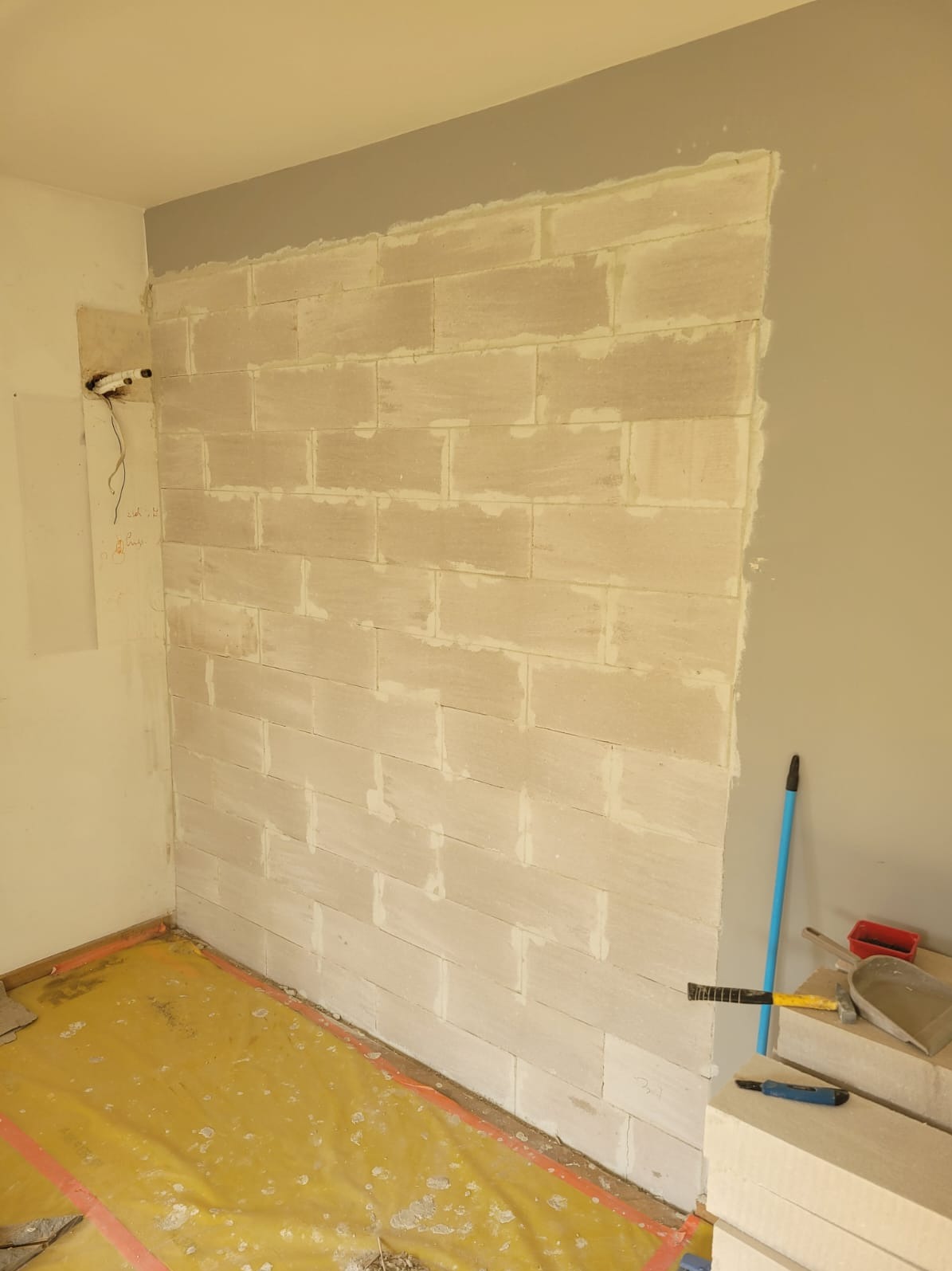 Ściana z bloczków z betonu komórkowego połączonych zaprawą, widoczne narzędzia murarskie, folia ochronna na podłodze. Fragment pomieszczenia w trakcie prac budowlanych.