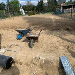 Przygotowany teren pod trawnik w Żywcu, z widocznymi dwoma taczkami, walcem i kratką drenażową, gotowy do dalszych prac.