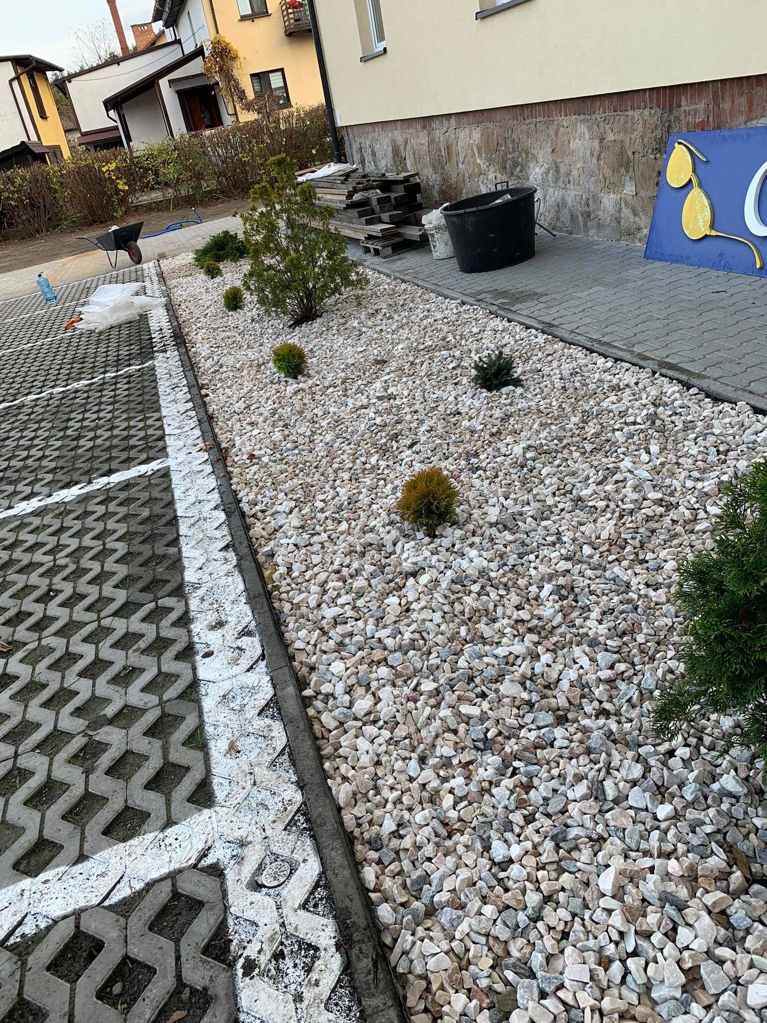 Nowo nasadzone tuje i inne krzewy ozdobne na rabacie wysypanej białym grysem, oddzielone od parkingu z ażurowych płyt betonowych obrzeżem, widoczne narzędzia i wiadro.