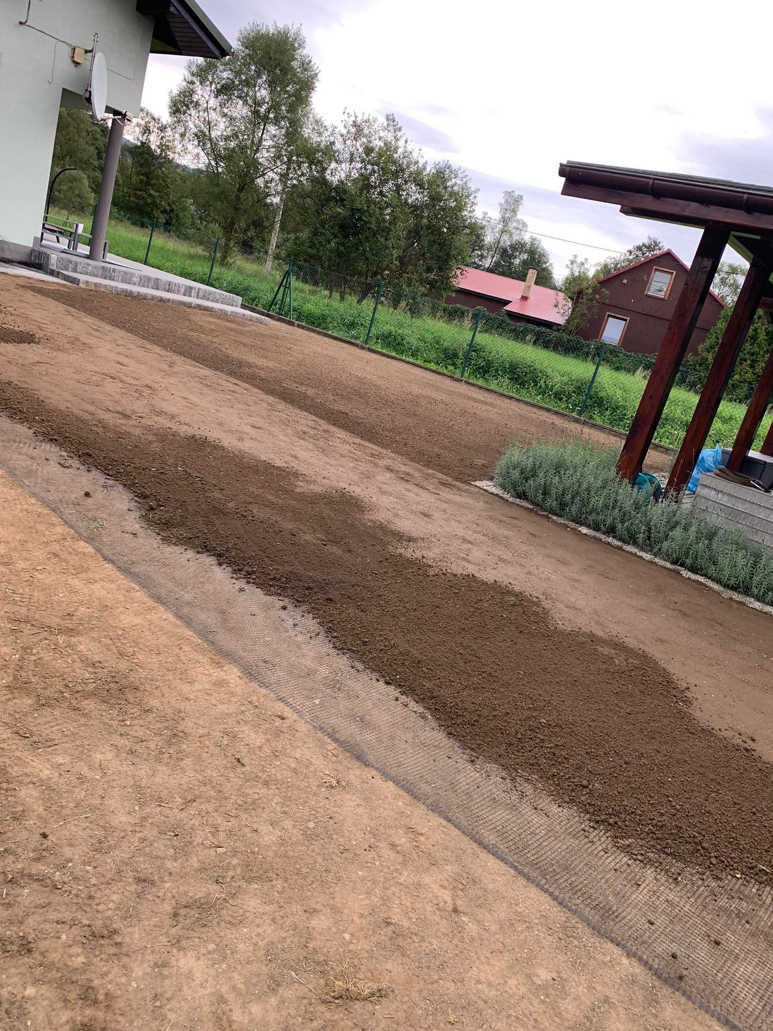 Przygotowanie gruntu pod trawnik z rolki, widoczna siatka wzmacniająca wzdłuż krawędzi, świeżo nawieziona ziemia, w tle dom i altana.